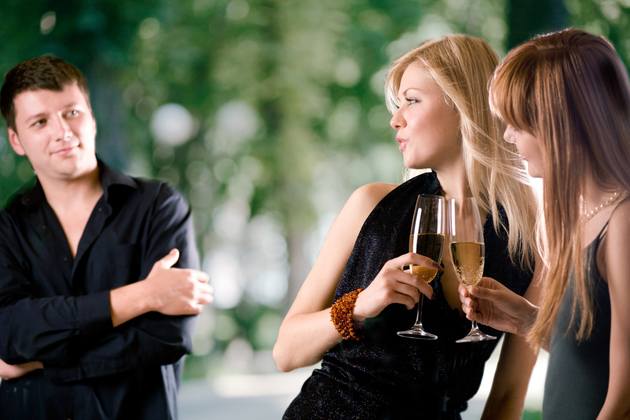 Two women holding glasses with champagne and laughing, young man looking at them and smiling, outdoors, focus on women